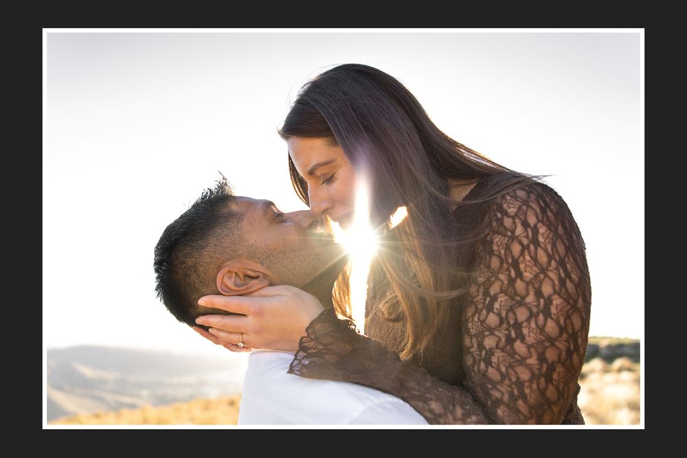 A couple shares a romantic kiss outdoors with golden sunlight glowing between them in a scenic landscape.