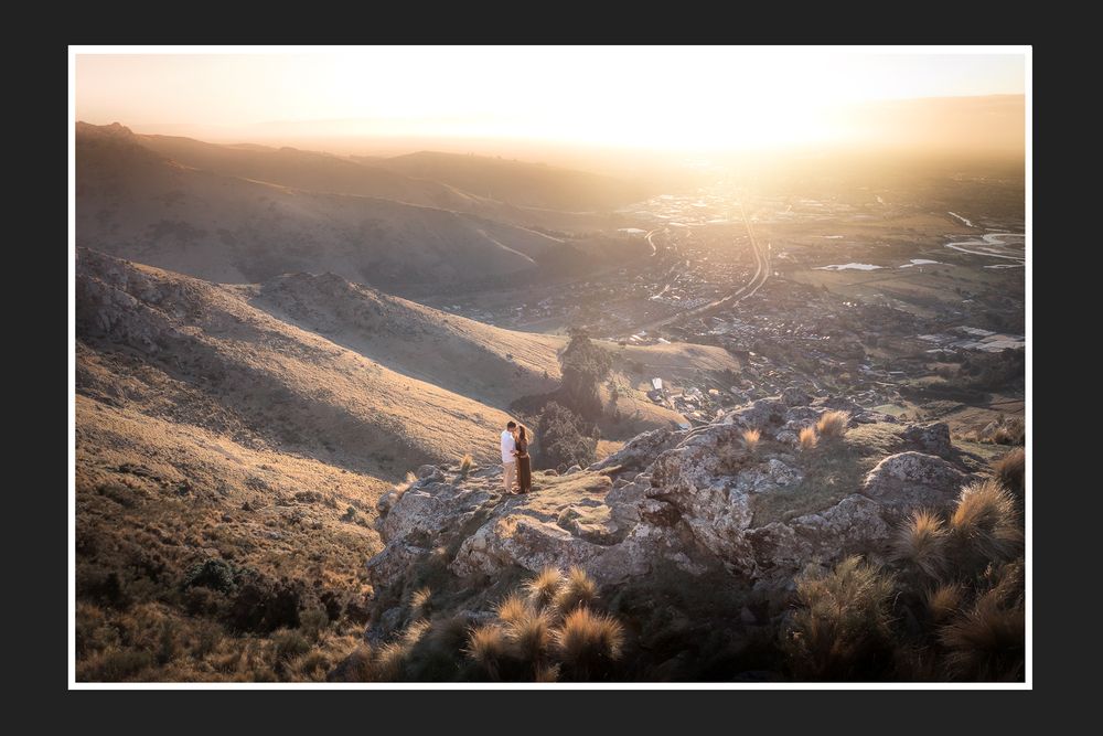 Two hikers stand on rocky outcrop overlooking golden sunlit valley and rolling hills at sunset.