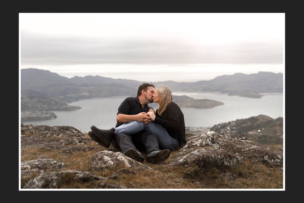Couple shares intimate moment while sitting on rocky overlook with lake view below.