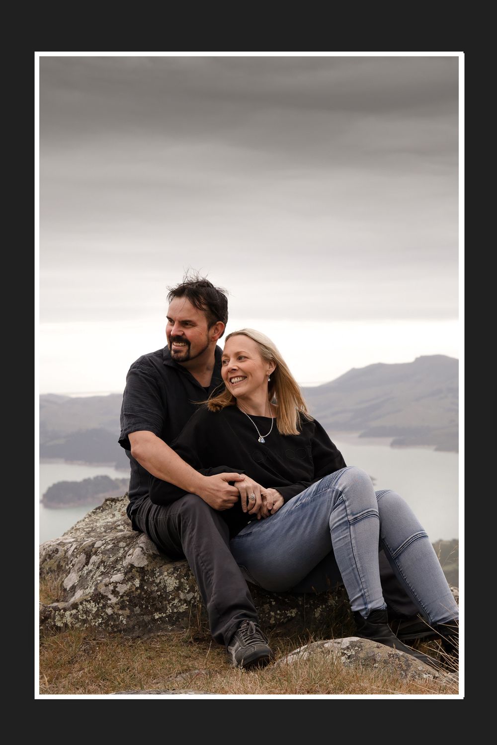 Candid couple laughing while sitting on rocky overlook with scenic mountain vista.