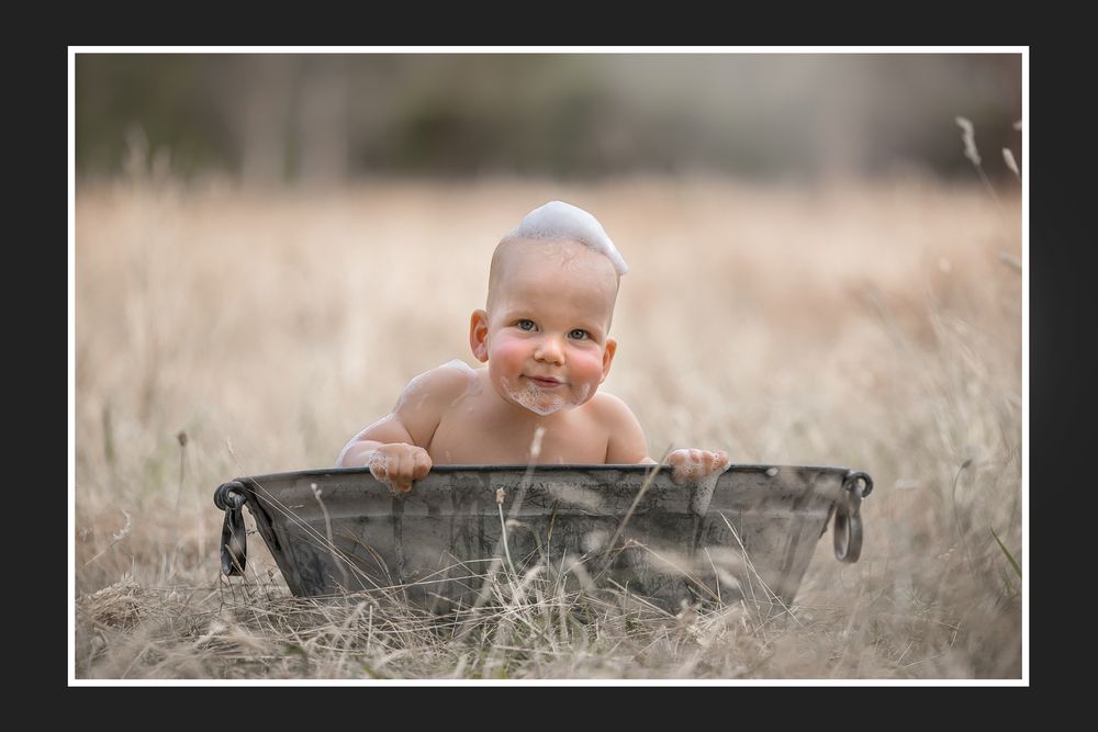 A baby lies in a rustic metal tub outdoors in a field of tall grass during a lifestyle portrait session.