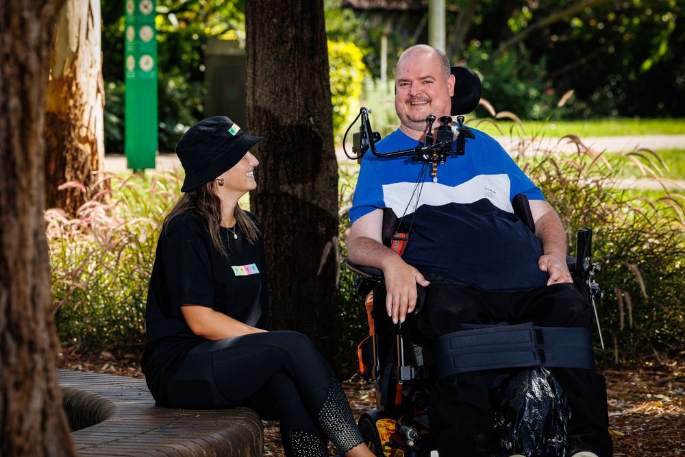 A support worker and client chat while sitting outdoors in a sunny garden setting.