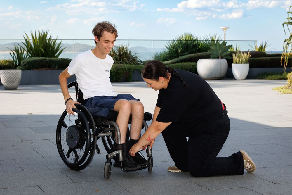A healthcare worker assists a wheelchair user on an outdoor terrace with planters.
