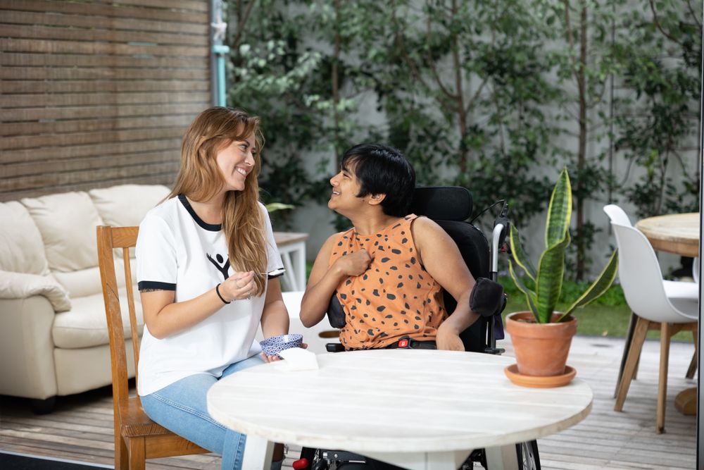 Two women having an animated conversation at a white table with a plant in the background.