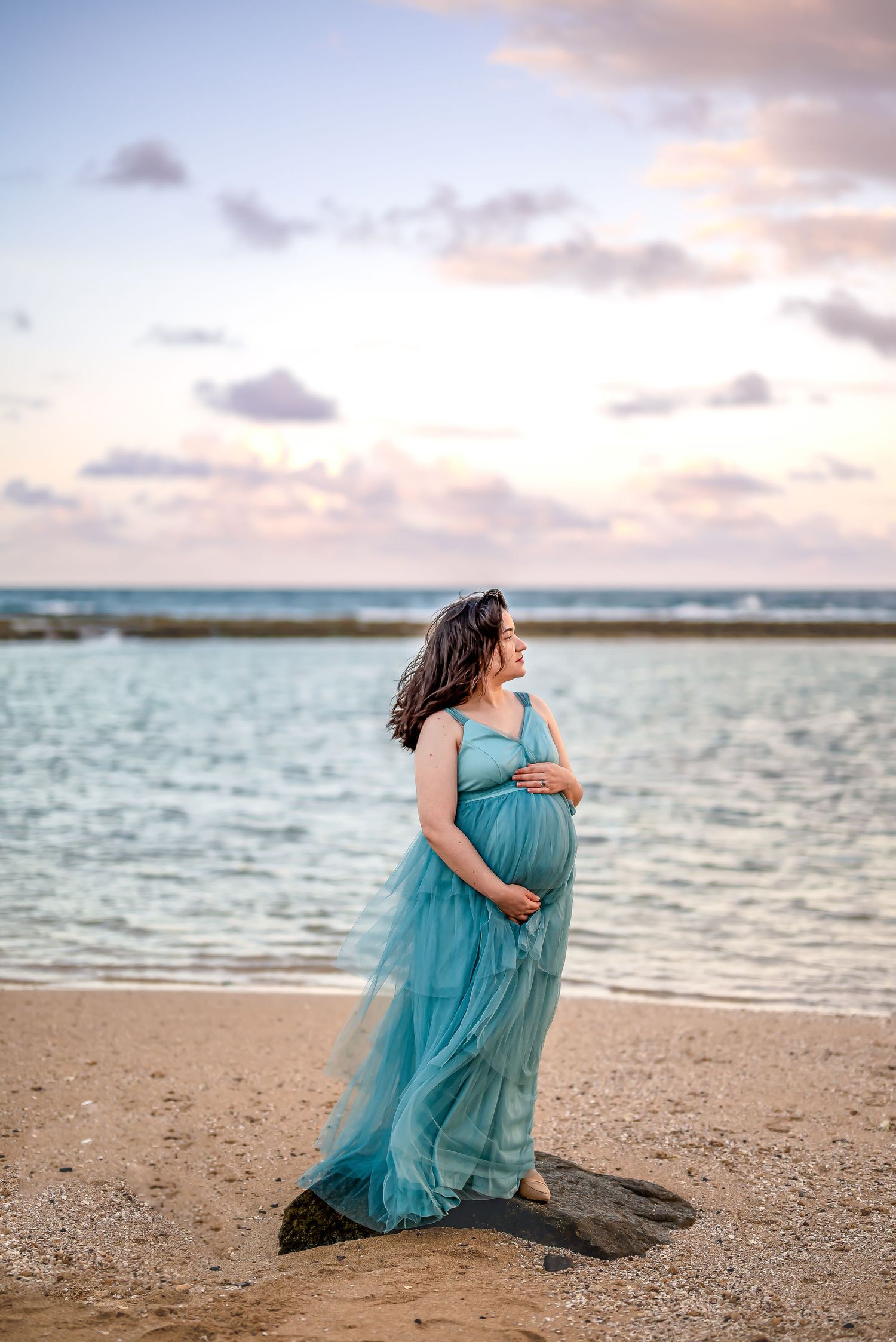 A pregnant woman in a flowing turquoise dress poses gracefully on a beach at sunset with ocean waves in the background.