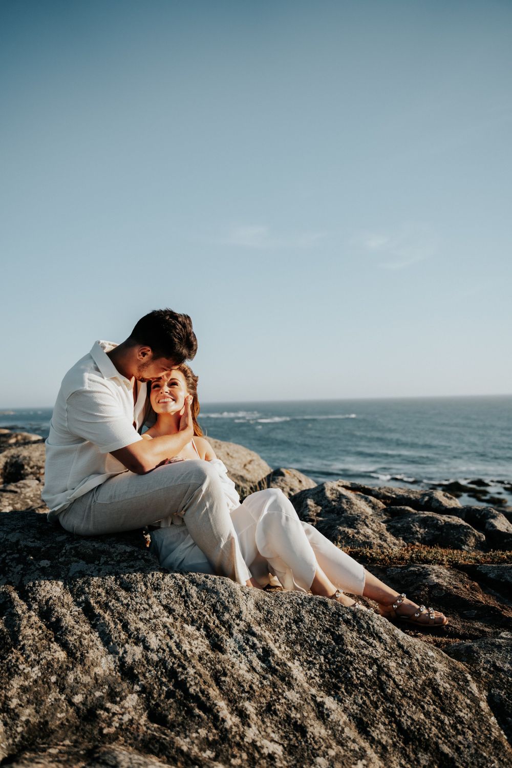 Couple sits together on coastal rocks overlooking ocean during sunset.