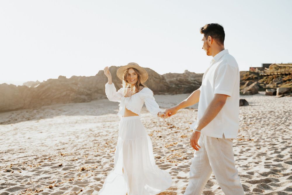 Couple in matching white outfits dance together on beach with cliffs in background.