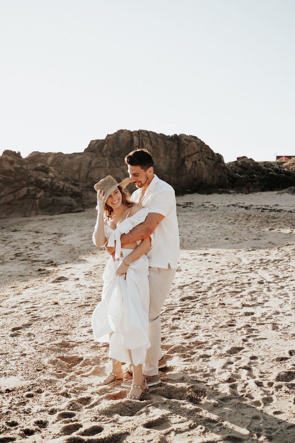Couple shares intimate moment while standing together on sandy beach with rocky backdrop.