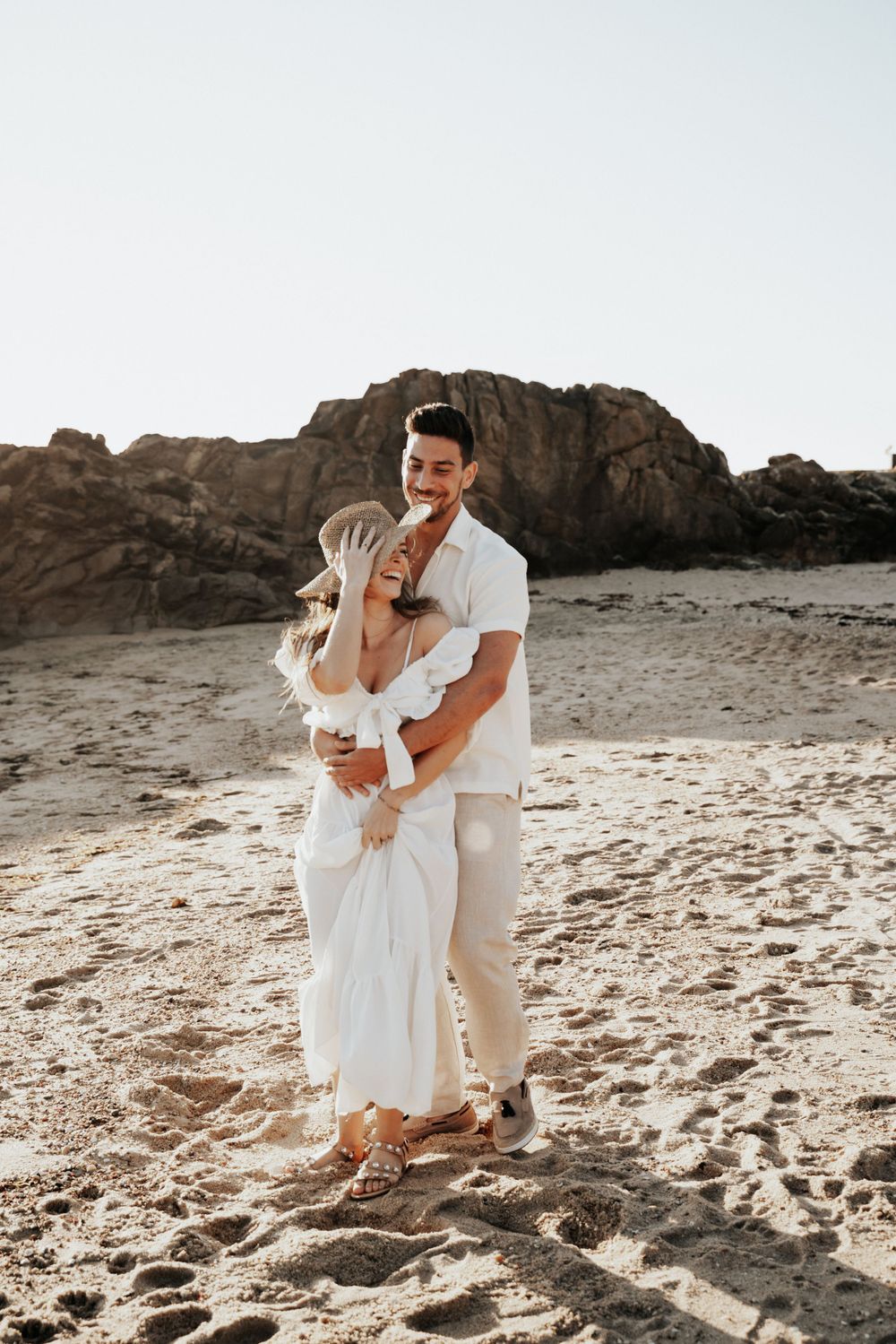 Romantic couple in white outfits embrace on a rocky beach during golden hour.