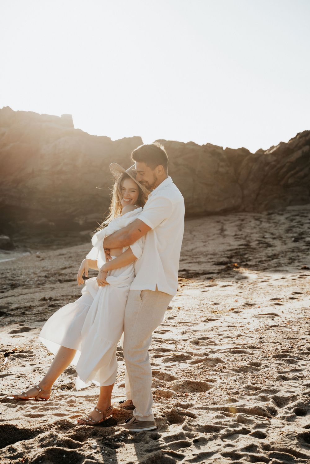 A couple in white clothing embraces on a sandy beach with rocky cliffs behind them.