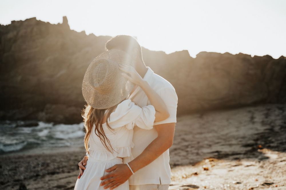 A couple in white shares a romantic moment on a rocky beach at sunset.