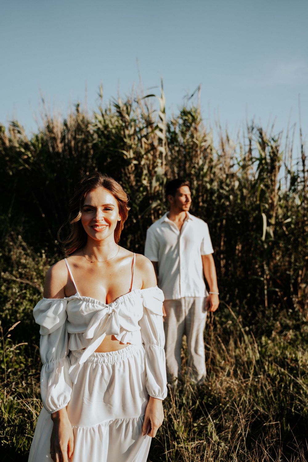 A carefree couple posing in a field of tall grass wearing white summer clothing.