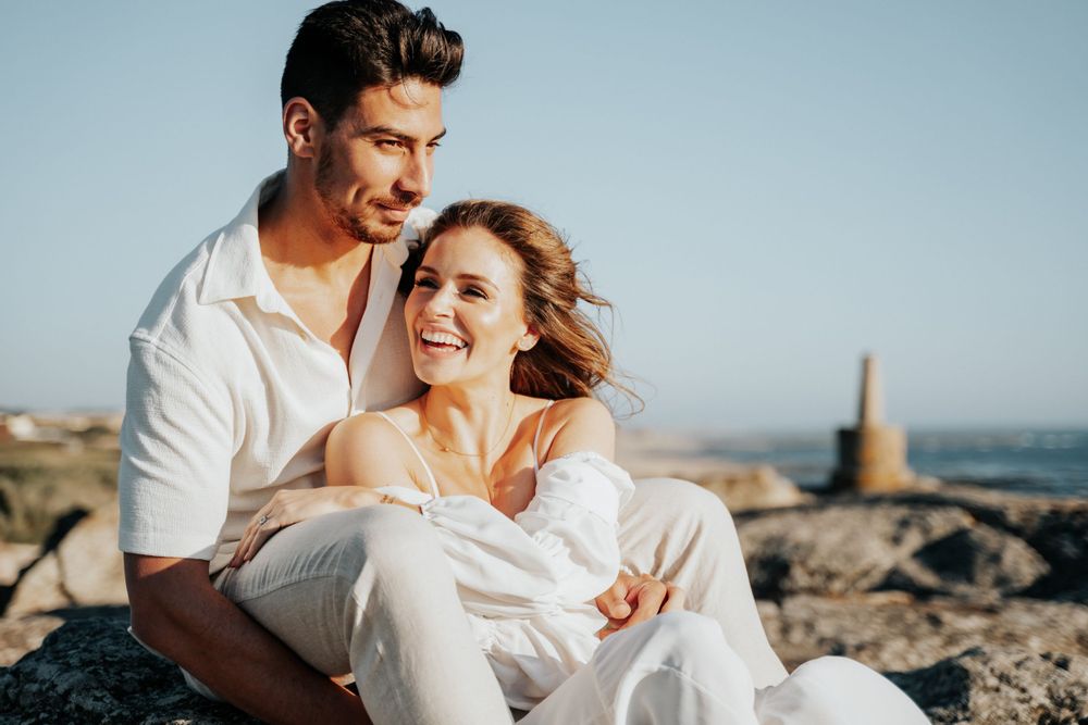 A playful couple laughing together while sitting on coastal rocks by the ocean.