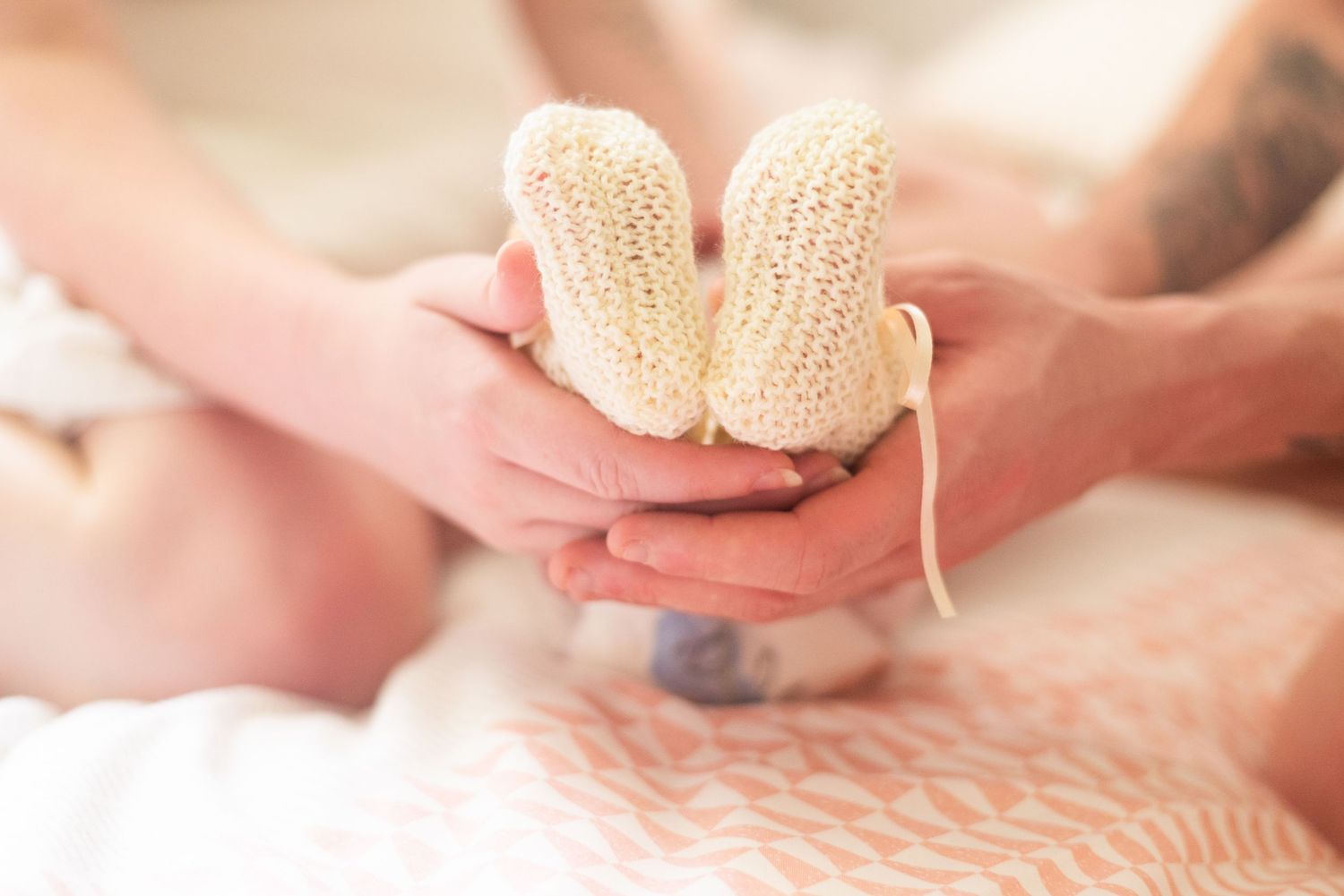 Small knitted baby booties held gently in hands against a soft pink background.