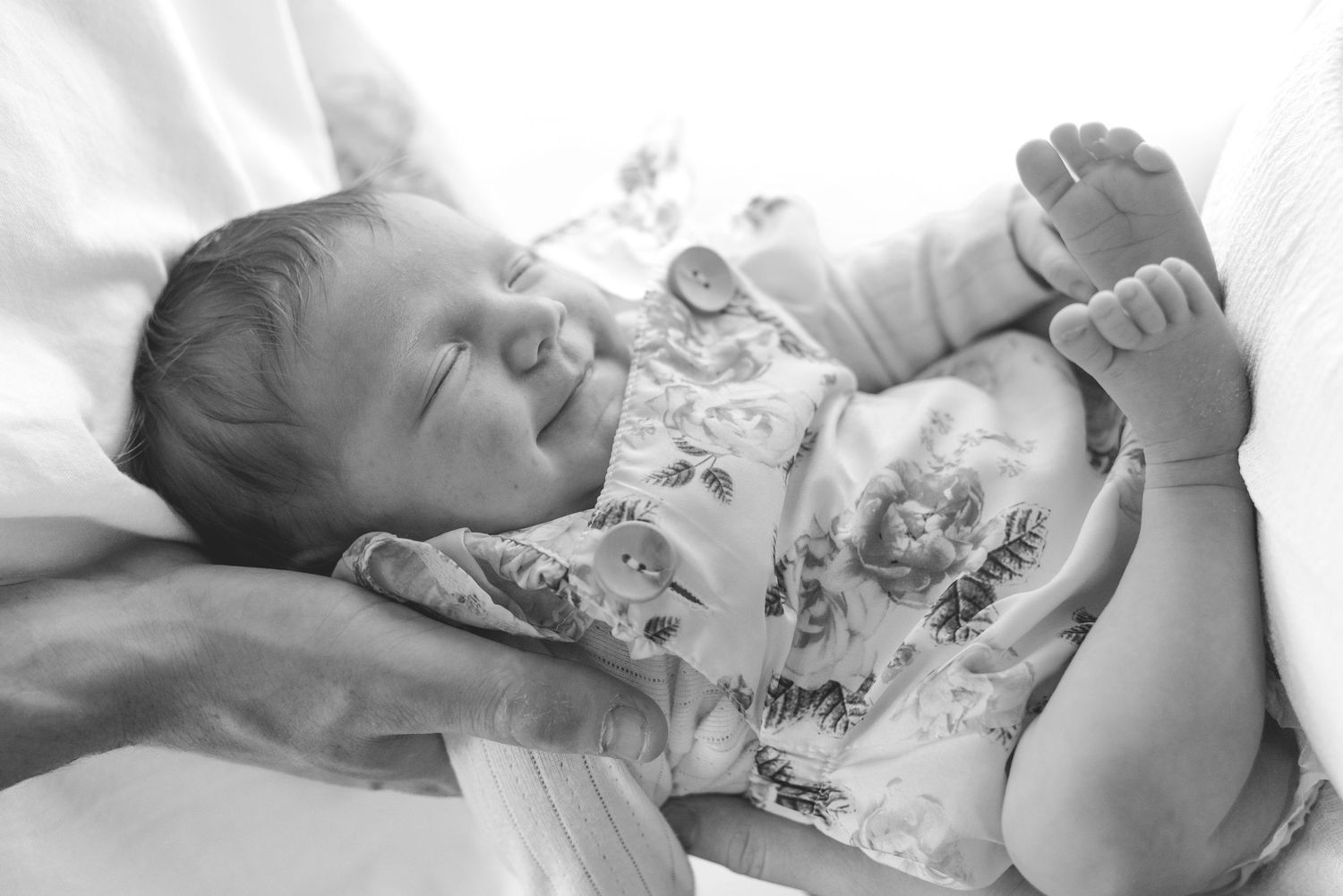 Black and white portrait of a smiling baby lying on white bedding.