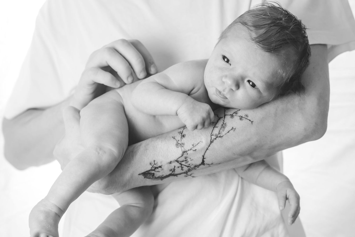 Black and white portrait of newborn baby resting on tattooed arms.