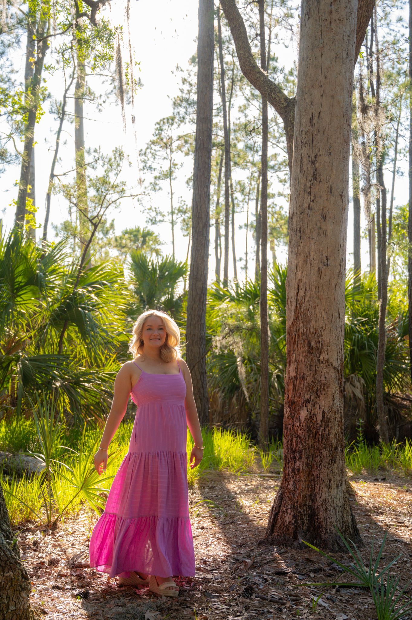A woman in a flowing pink dress stands among tall pine trees and palm fronds in dappled sunlight.