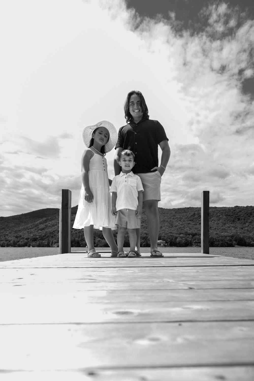 Black and white photo of people standing on a wooden dock overlooking water and mountains.