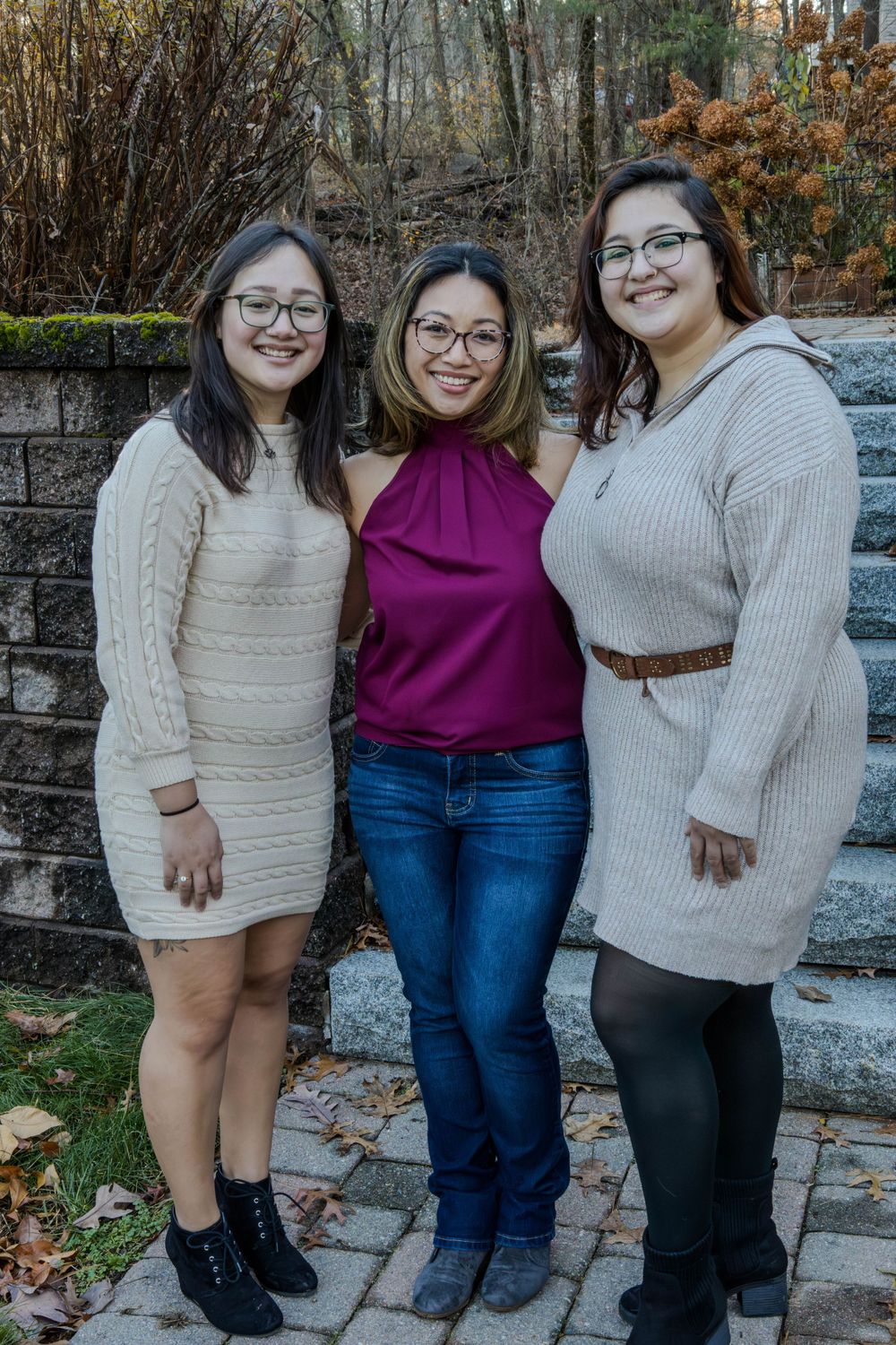 Three friends in casual sweaters and jeans pose together on stone steps during autumn.
