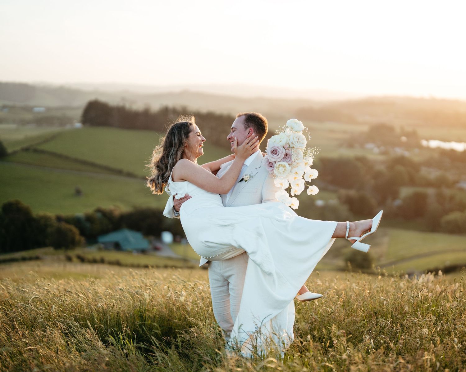 A couple celebrates their wedding day with a romantic pose in a hilltop field at sunset at Kauri bay wedding venue in Auckland.
