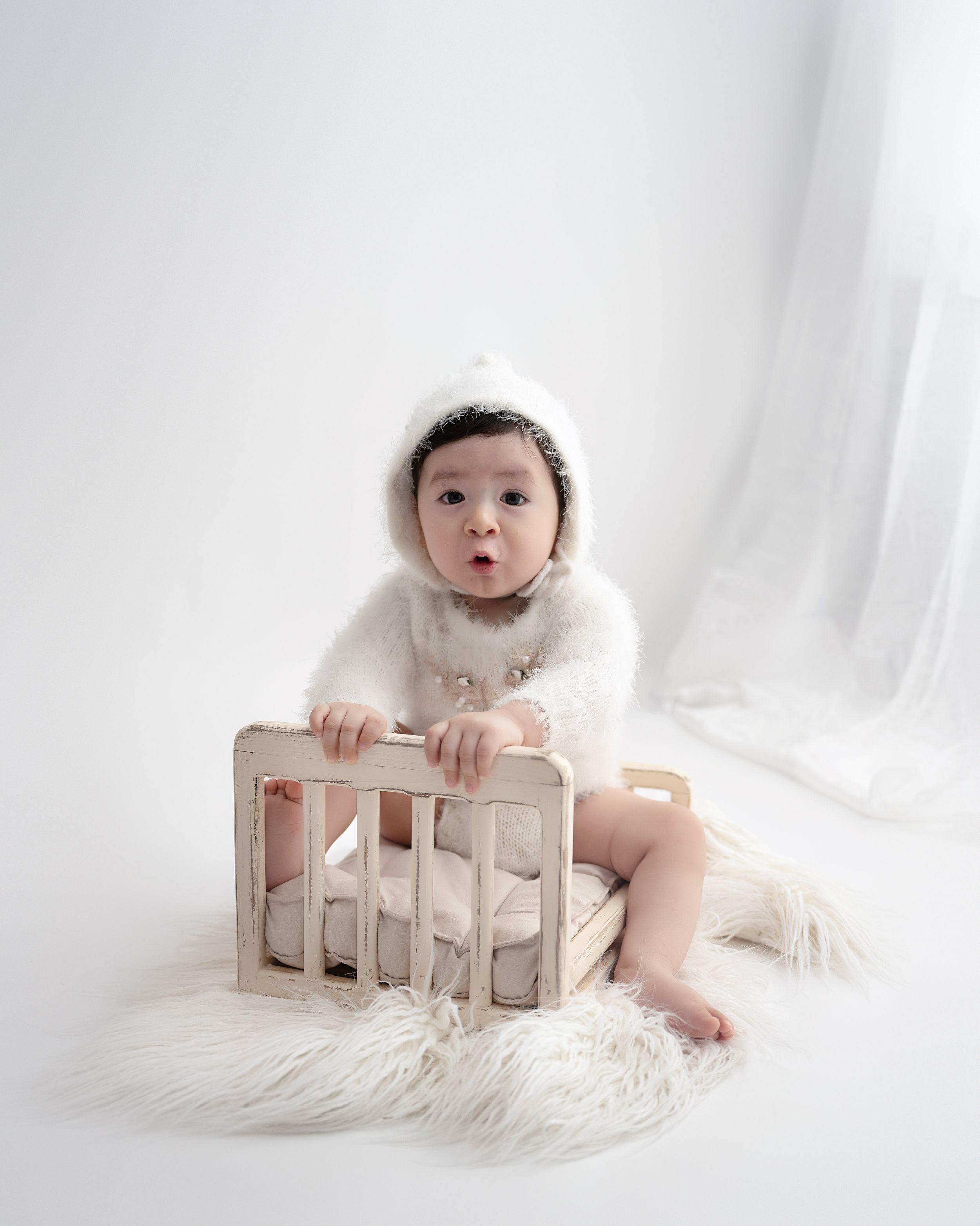 Minimalist studio photo of baby in white sweater sitting against wooden crate on faux fur rug with white backdrop.