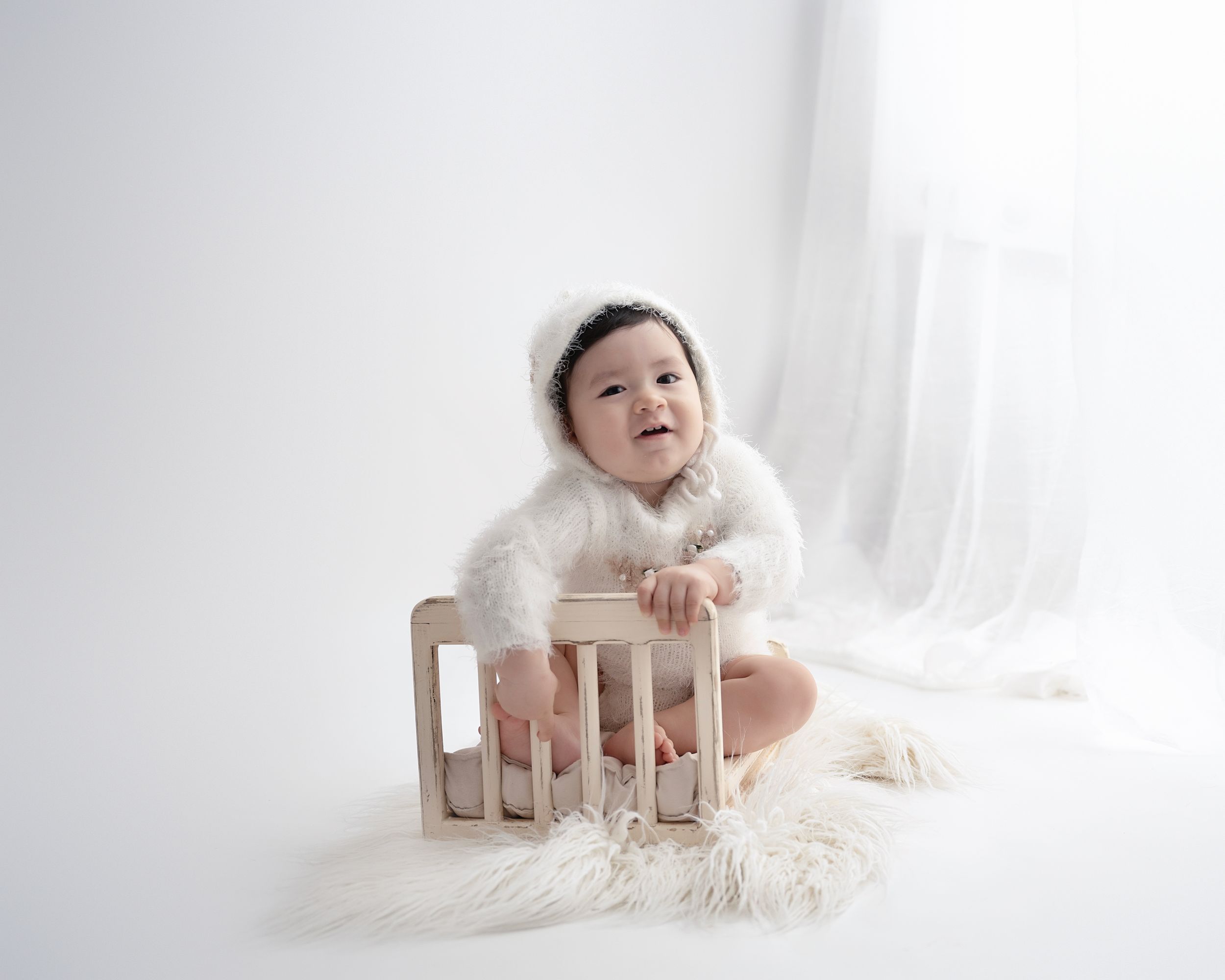 Artistic portrait of baby in cozy white sweater posing with wooden crate on furry white background.
