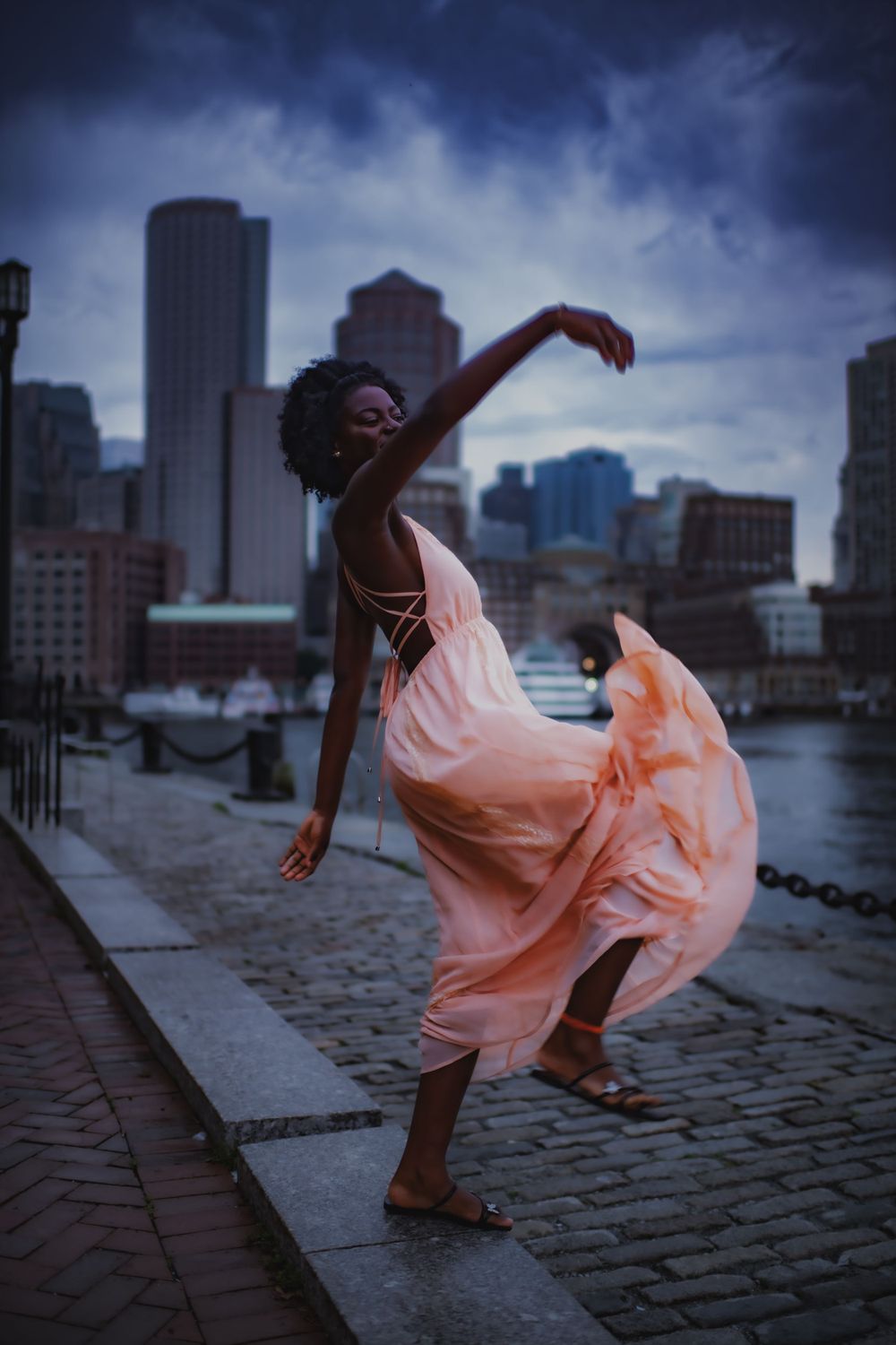 Dramatic dance pose in a flowing peach dress against a moody city skyline at dusk.