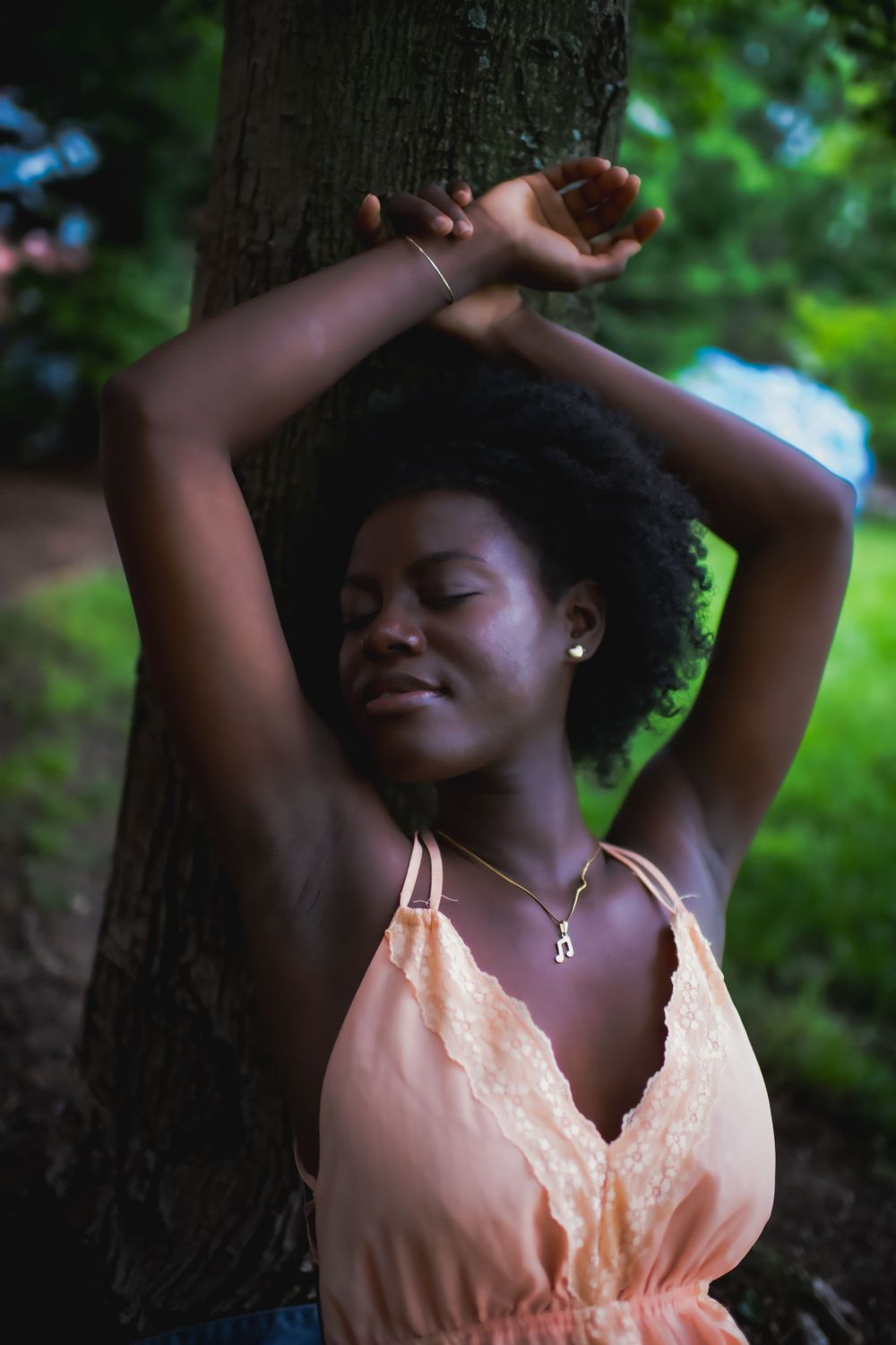 Peaceful pose in a peach-colored dress against lush green foliage and a tree trunk.