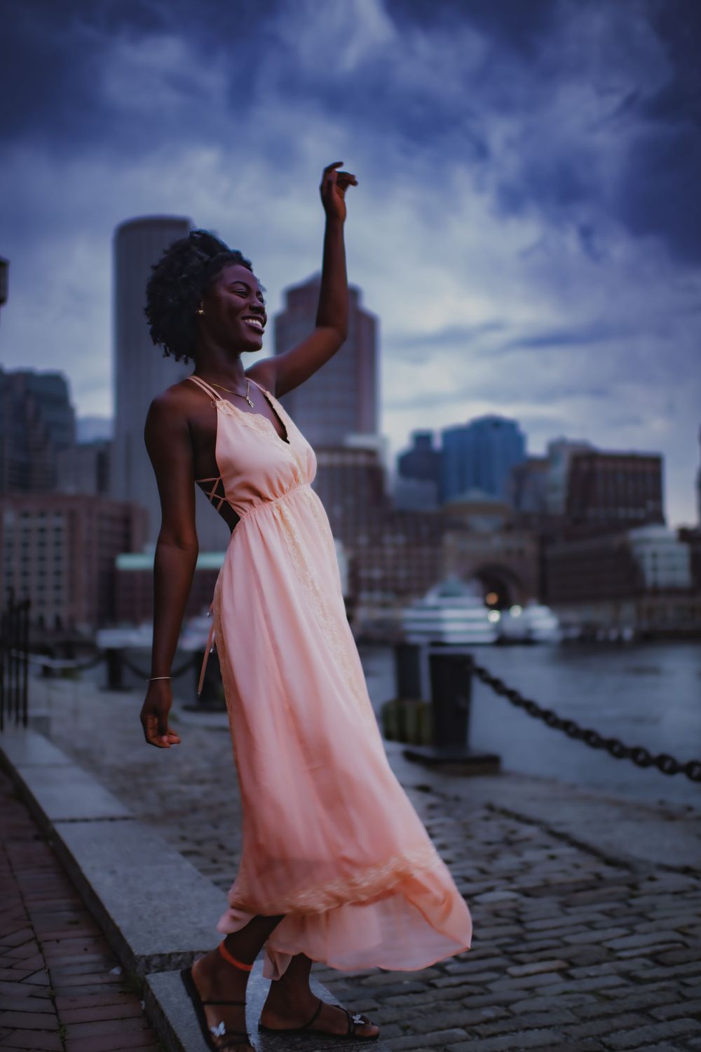 Joyful dance pose in flowing pink dress against city harbor backdrop.