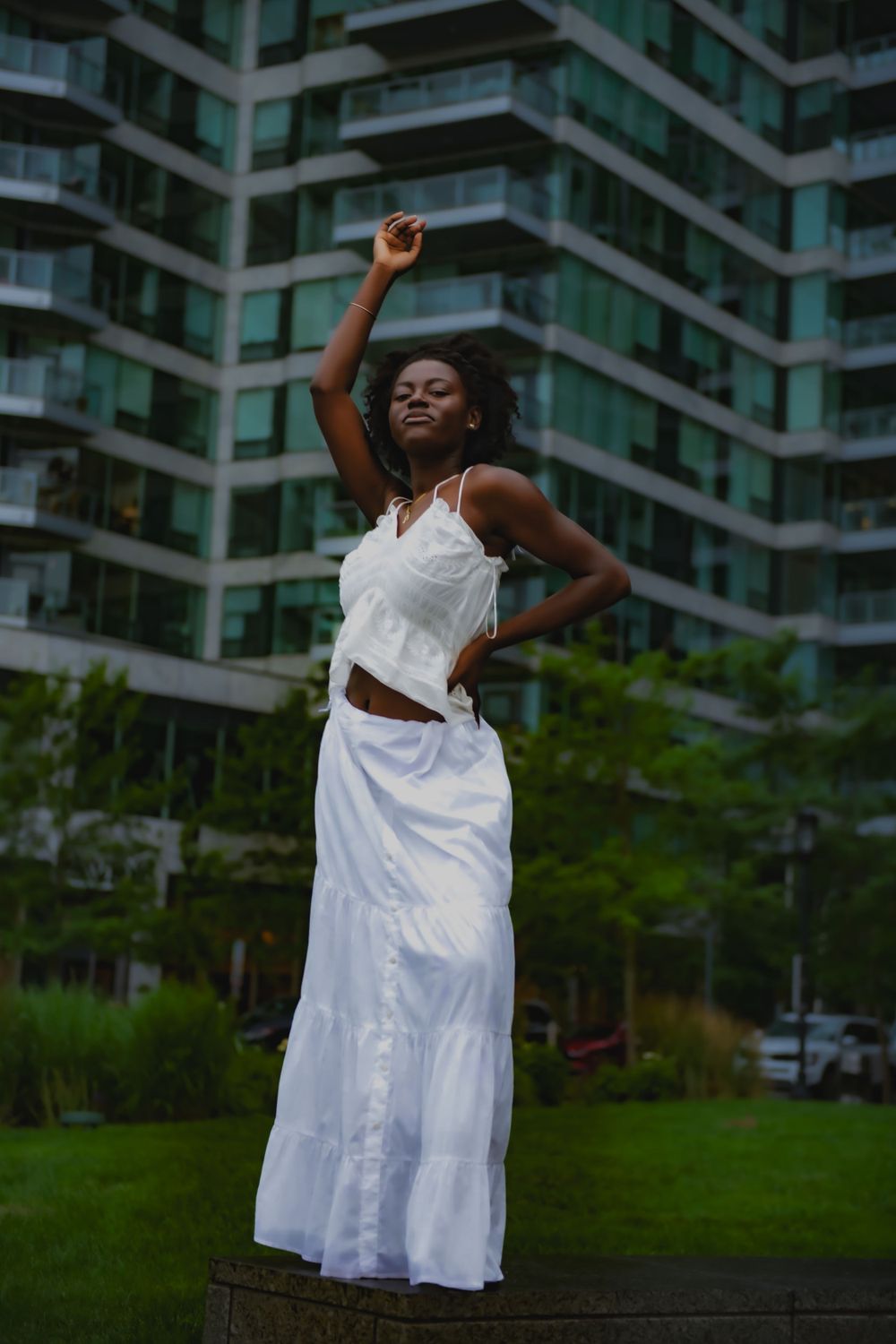 A graceful figure in a flowing white dress poses elegantly against a modern apartment building backdrop.
