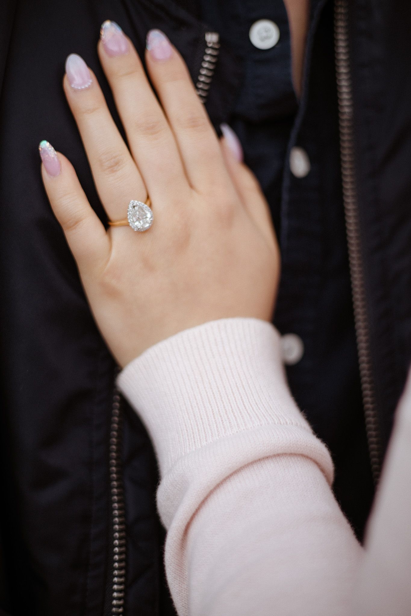Hand wearing a large teardrop-shaped diamond engagement ring with french manicured nails and pink sweater sleeve.