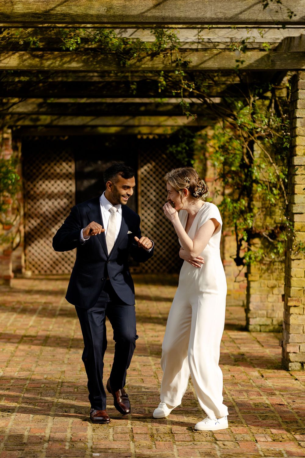 A couple in formal attire dancing and laughing together under a rustic garden pergola.