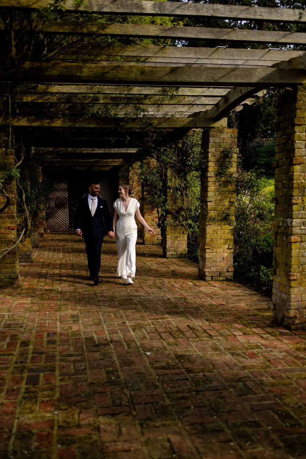 A couple in wedding attire walking through a brick-paved garden pergola with climbing vines.