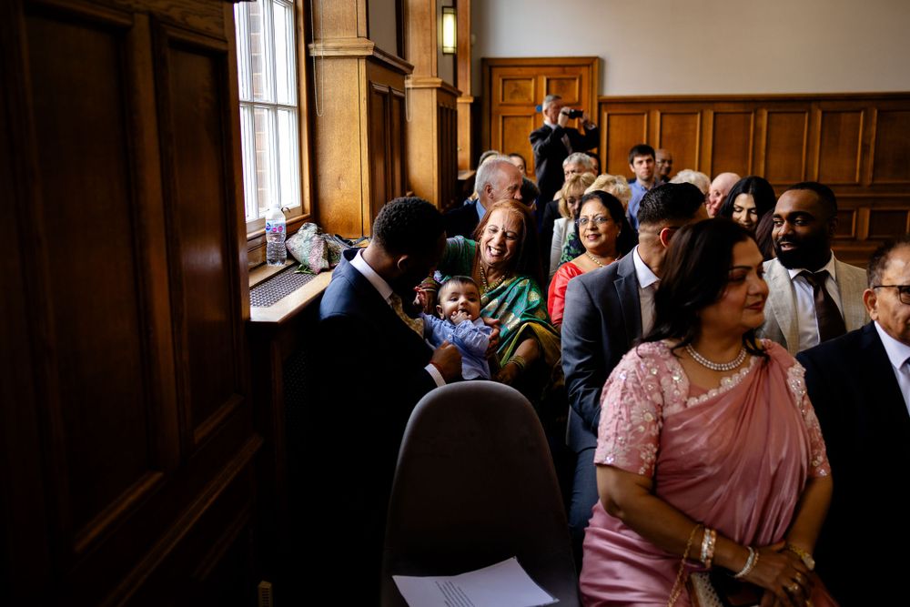 Diverse group of wedding guests gathered by window in wood-paneled room.