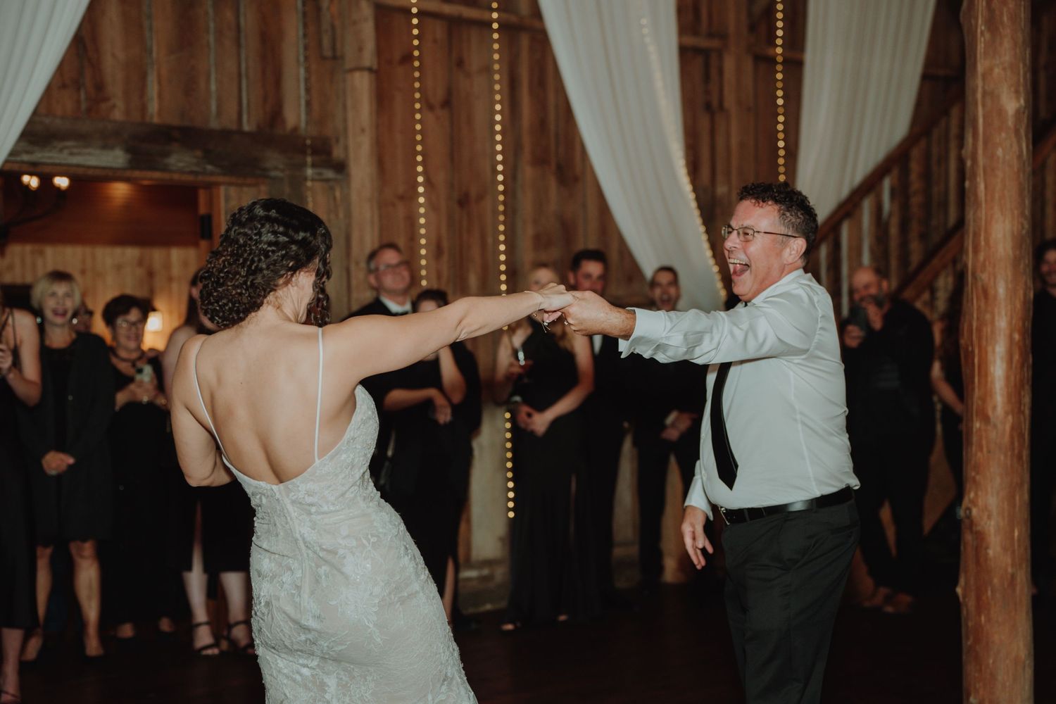 Bride in backless gown dances with guest at lively rustic barn wedding reception.