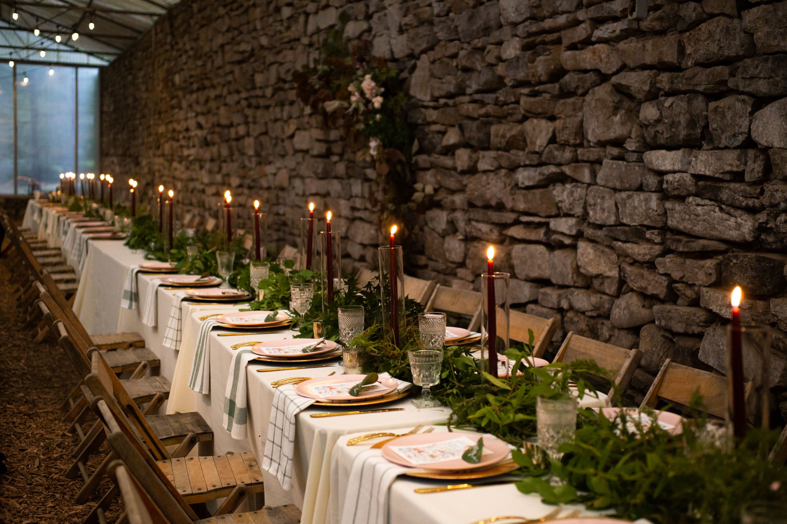 Elegant wedding reception table with candles and greenery against rustic stone wall.