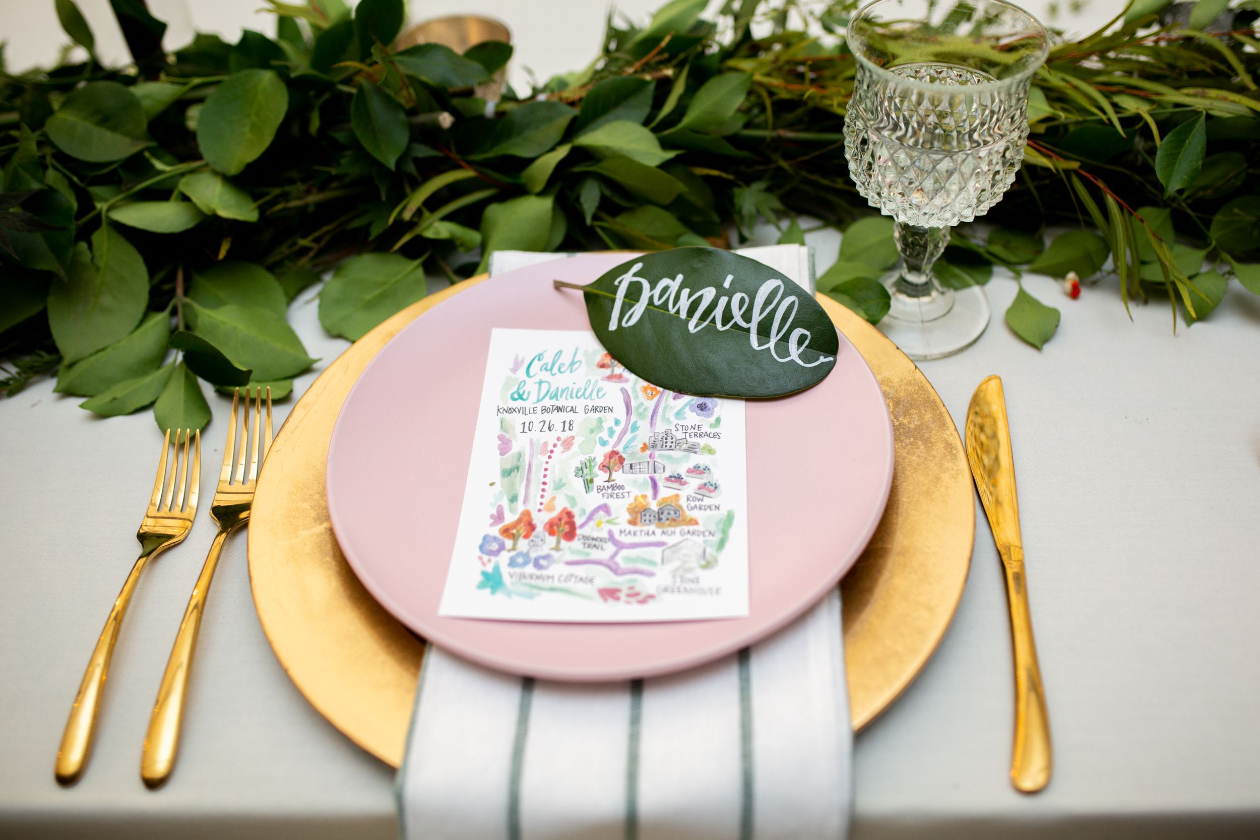 Wedding place setting with pink plate, gold charger, and greenery detail.