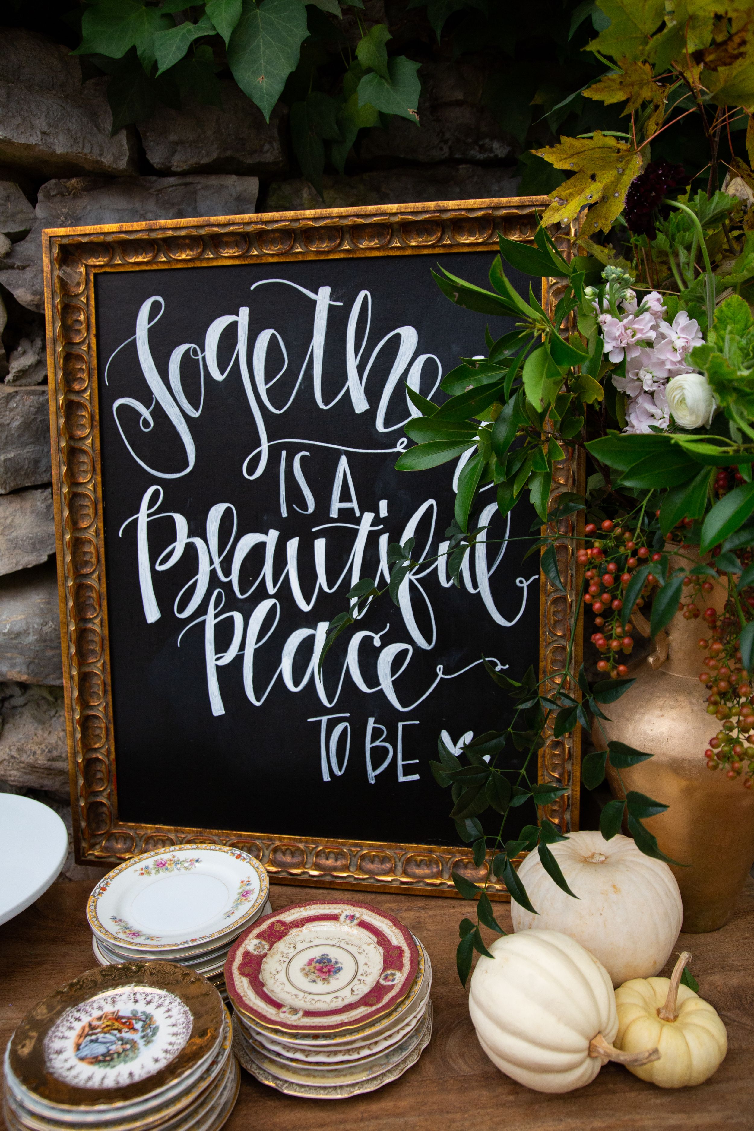 Framed chalkboard sign with white calligraphy surrounded by white pumpkins and vintage plates.