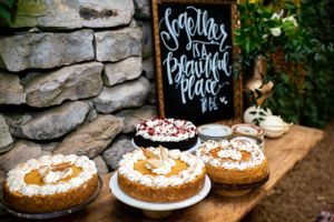Three decorated cakes displayed on wooden table with chalkboard sign and stone wall backdrop.