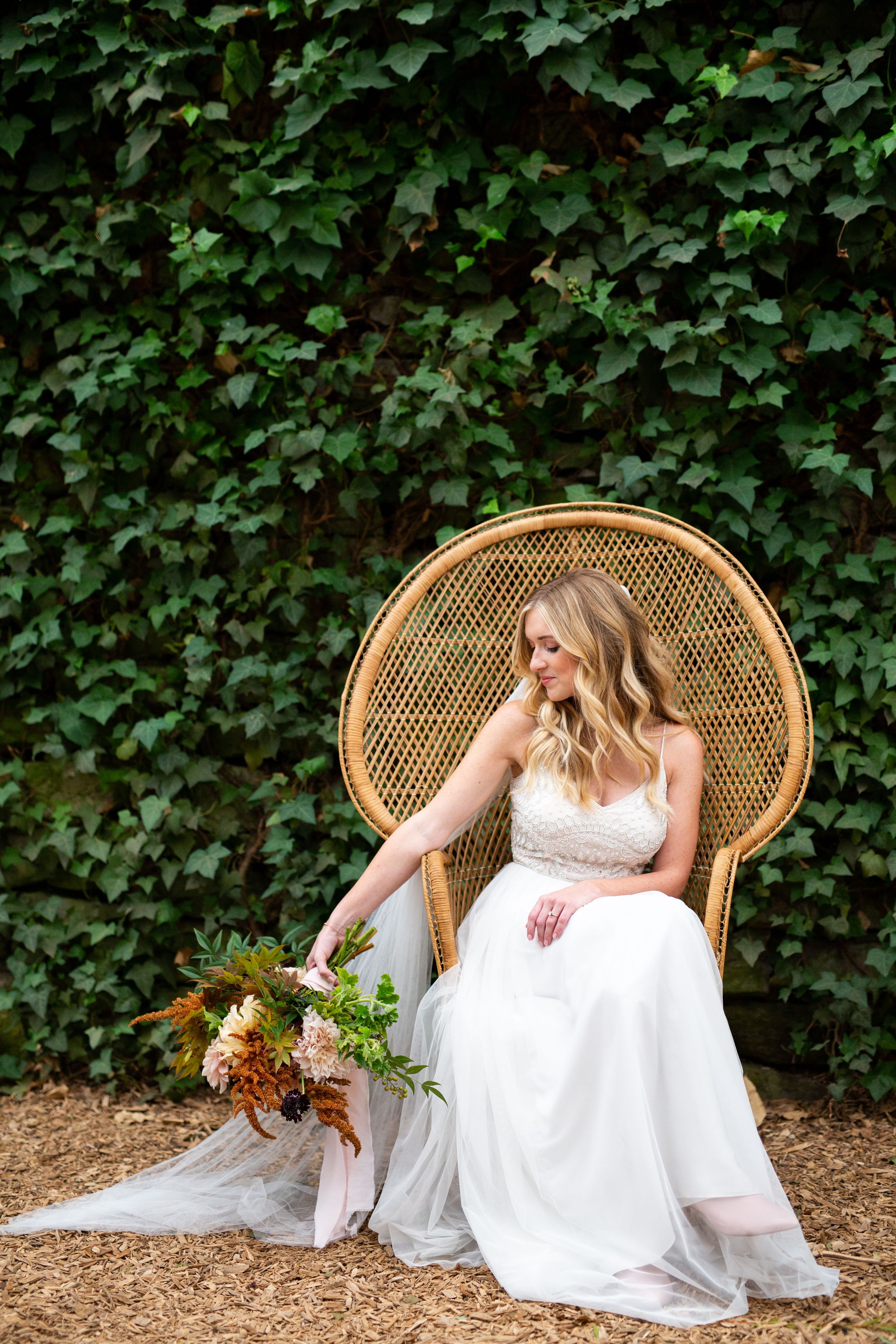 A bride in white dress sits in a peacock chair against an ivy wall with a fall bouquet.