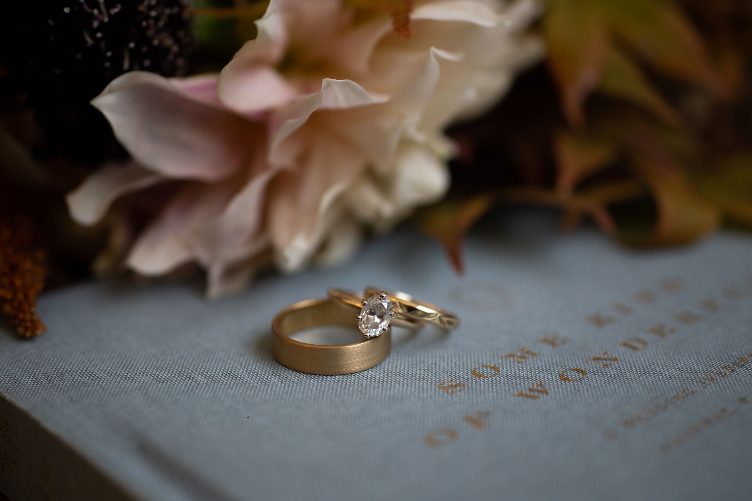 Two gold wedding rings photographed with soft pink flowers and subtle text detail.