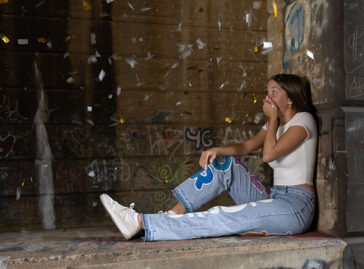 Person sitting against graffiti wall wearing jeans and white sneakers in urban setting.