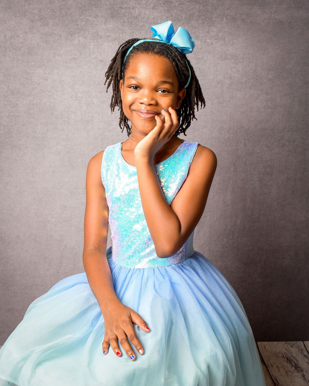 Young girl in a light blue princess dress and bow posing against grey backdrop with playful smile.