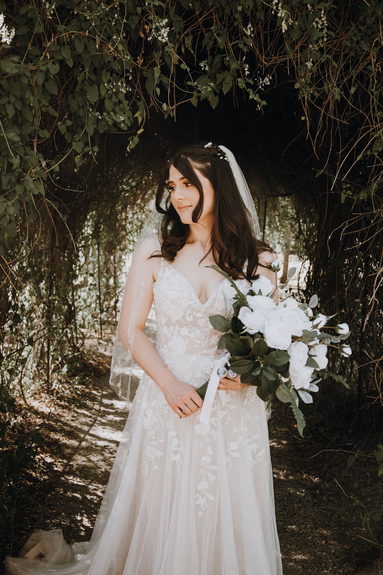 Bride in lace wedding gown holding white bouquet standing under green vine archway.