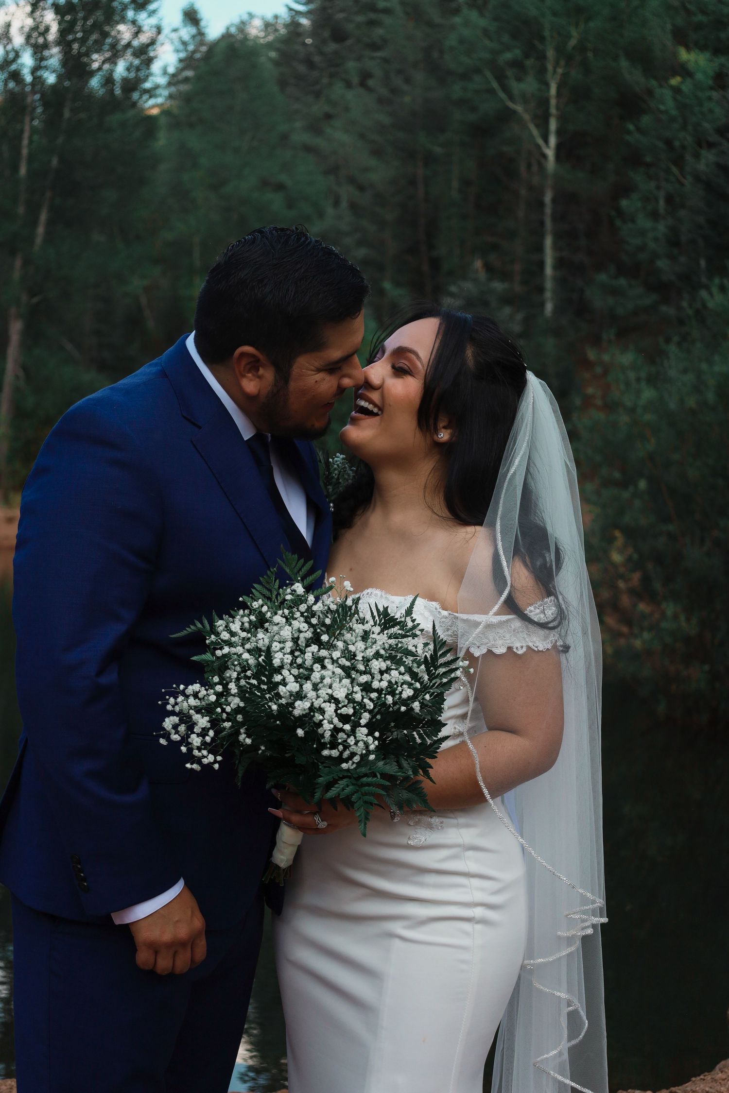 Groom in blue suit kissing bride on cheek, bride in off-shoulder gown holding baby's breath bouquet.