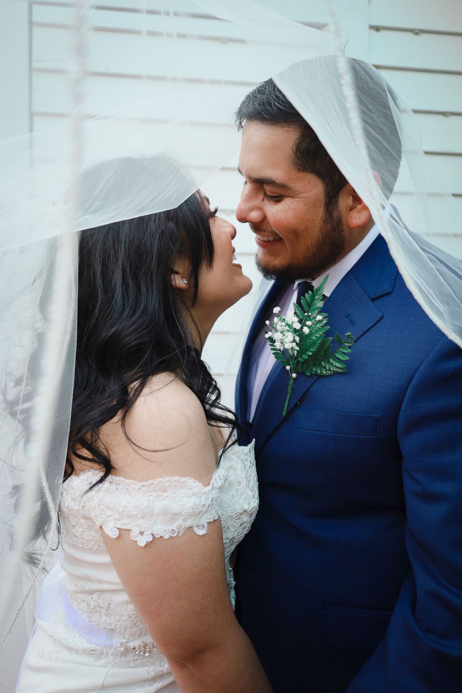 Bride and groom sharing romantic moment under wedding veil, groom wearing blue suit with boutonniere.