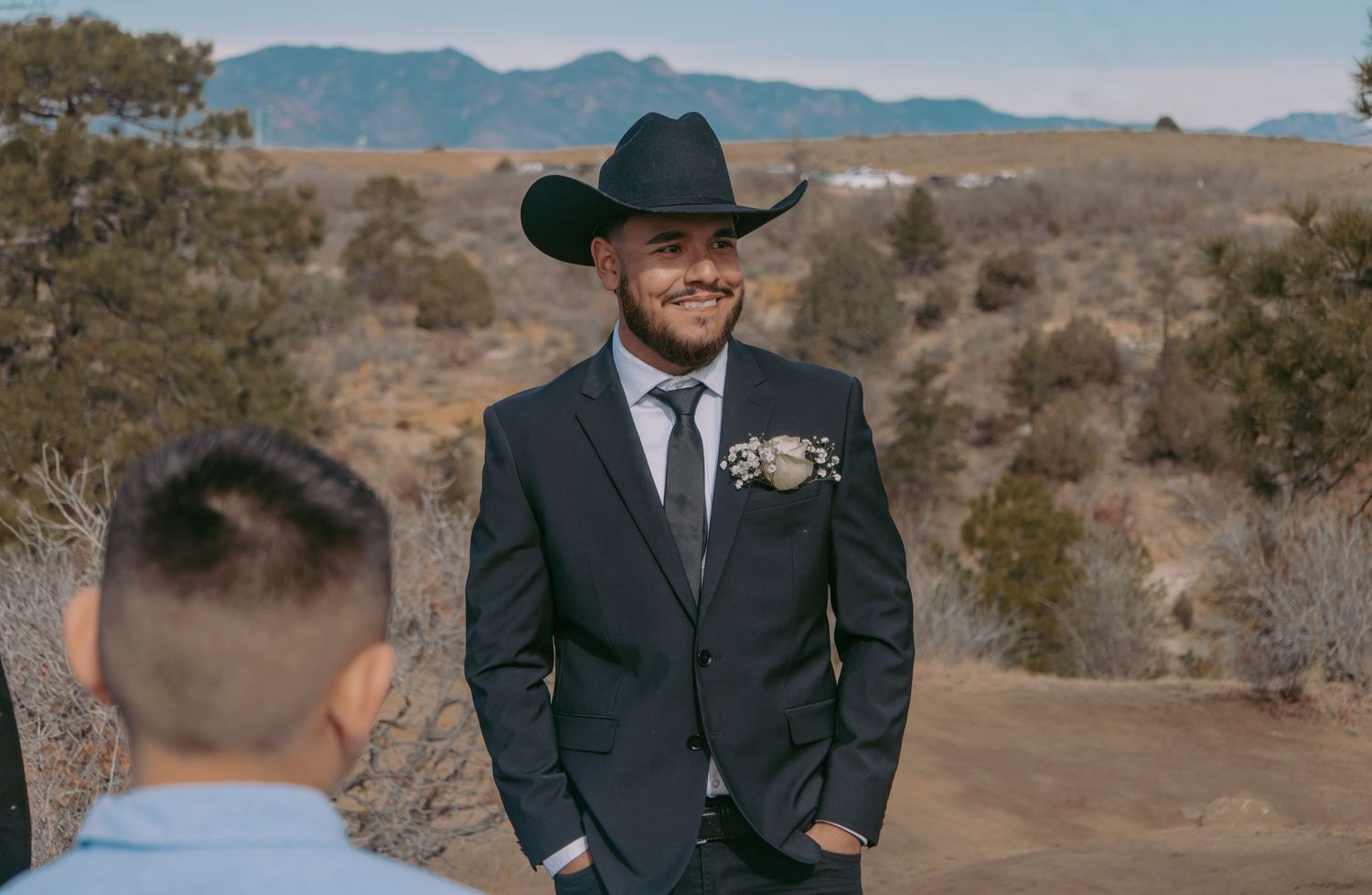 Smiling groom in black cowboy hat and suit with boutonniere at outdoor desert wedding ceremony.