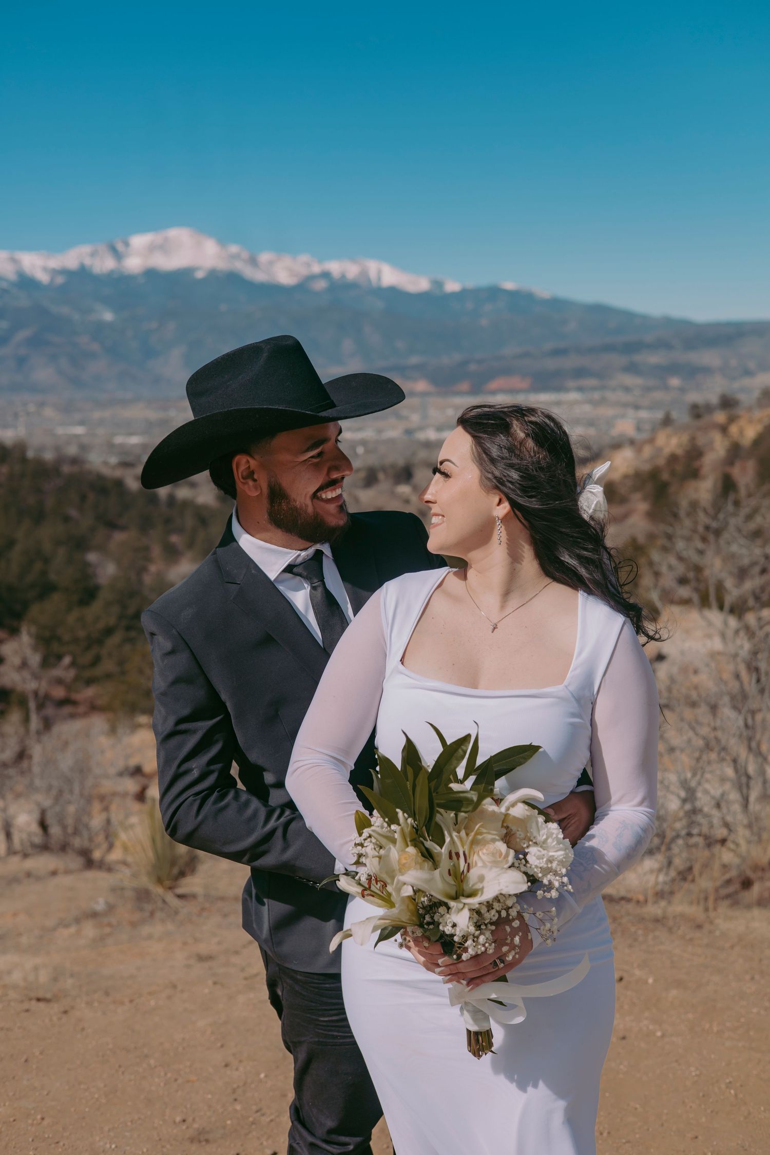 Bride and groom laughing together outdoors, groom in black cowboy hat, bride holding white bouquet with mountains behind.