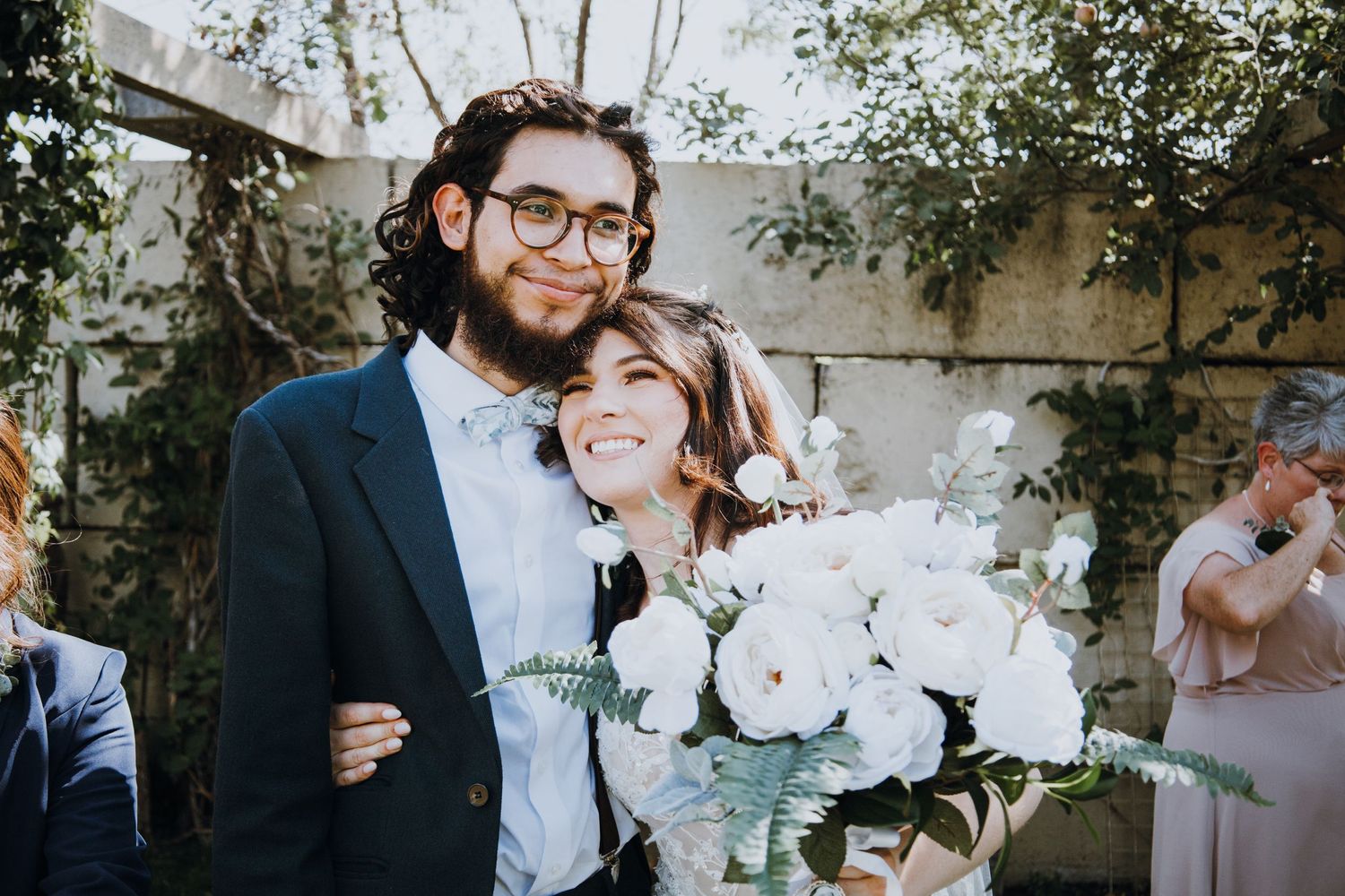 Happy couple smiling after wedding ceremony, groom in navy suit, bride holding white peony bouquet.