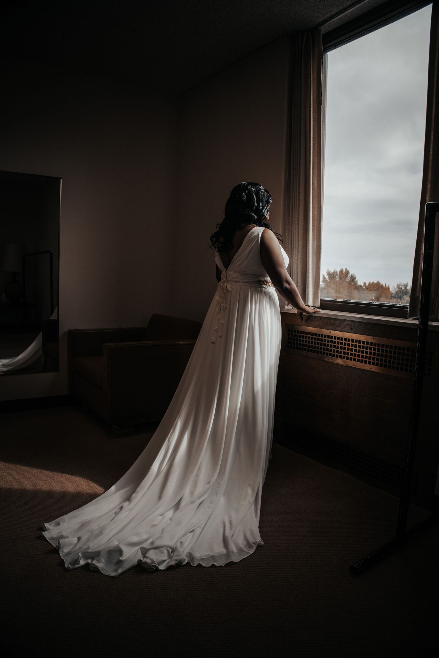 Bride in elegant white wedding gown with train standing at window in dramatic lighting.